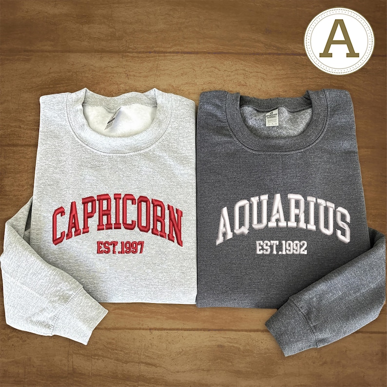 May include: Two grey sweatshirts with embroidered text. One is light grey with "CAPRICORN EST.1997" in red. The other is dark grey with "AQUARIUS EST.1992" in white. Both have a crew neck and long sleeves.