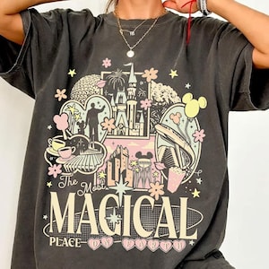 以下が含まれることがあります： お城、ミッキーマウス、その他のテーマパーク要素のカラフルなグラフィックが特徴の濃い灰色のTシャツ。「The Most Magical Place on Earth」の文字が下に表示されています。シャツは半袖で、クルーネックです。