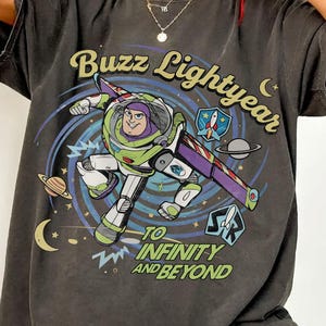 May include: Dark gray t-shirt featuring a graphic of Buzz Lightyear from Toy Story. The design includes Buzz in his spacesuit, the words "Buzz Lightyear" and "To Infinity and Beyond", along with planets and stars.
