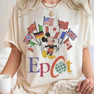 May include: Cream-colored t-shirt featuring Mickey Mouse holding flags from various countries. The word "Epcot" is printed in colorful letters below the graphic. The shirt has short sleeves and a relaxed fit.