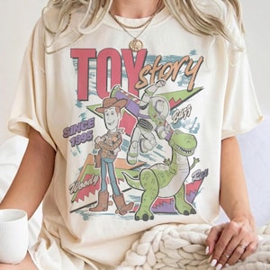 Puede incluir: Camiseta color crema con un gráfico de los personajes de Toy Story Woody, Buzz Lightyear y Rex. Las palabras "TOY STORY" y "SINCE 1995" están impresas encima y debajo de los personajes. La camiseta tiene mangas cortas.