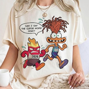 Könnte beinhalten: Cremefarbenes T-Shirt mit einer Cartoon-Grafik von zwei animierten Charakteren, die rennen. Einer ist rot und wütend, der andere orange und aufgeregt. Eine Sprechblase sagt: „CAN I SAY THE CURSE WORD NOW?“ Beide Charaktere tragen Lätzchen mit „2026“.