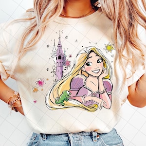 May include: Cream-colored sweatshirt featuring a cartoon illustration of a princess with long blonde hair, a purple dress, and a green chameleon. A purple tower, flowers, stars, and a sun are also depicted in the design.