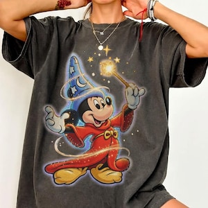 Könnte beinhalten: Dunkelgraues T-Shirt mit einer Grafik von Mickey Mouse als Zauberer, der einen leuchtenden Zauberstab hält. Mickey trägt einen roten Umhang und einen blauen Zaubererhut mit Sternen und Monden. Das Design umfasst goldene Sterne und Wirbel.