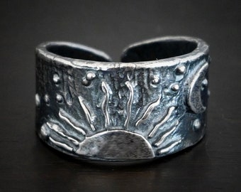 May include: A silver ring with a textured surface and a raised design of a sun and moon. The ring is a statement piece with a unique and eye-catching design.