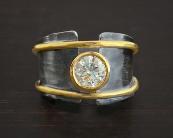 May include: A wide silver band ring with a large round diamond set in a bezel setting. The band is accented with three thin gold bands.