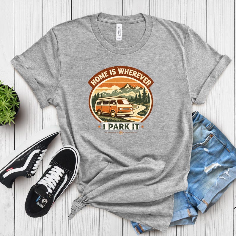 May include: A heather gray t-shirt featuring a circular graphic with a vintage camper van driving on a road through a mountain landscape. The text "HOME IS WHEREVER I PARK IT" is above and below the image. Black and white sneakers and denim shorts are also visible.