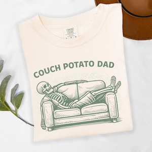 May include: Cream-colored t-shirt with the text "COUCH POTATO DAD" in green. Below the text is a green illustration of a skeleton relaxing on a couch. The shirt is folded and has a tag that says "COMFORT COLORS".