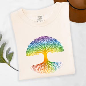 May include: Cream-colored t-shirt featuring a vibrant rainbow-colored tree design. The tree's branches and roots are depicted in a spectrum of colors, from red to purple. The shirt has a tag that says "COMFORT COLORS".