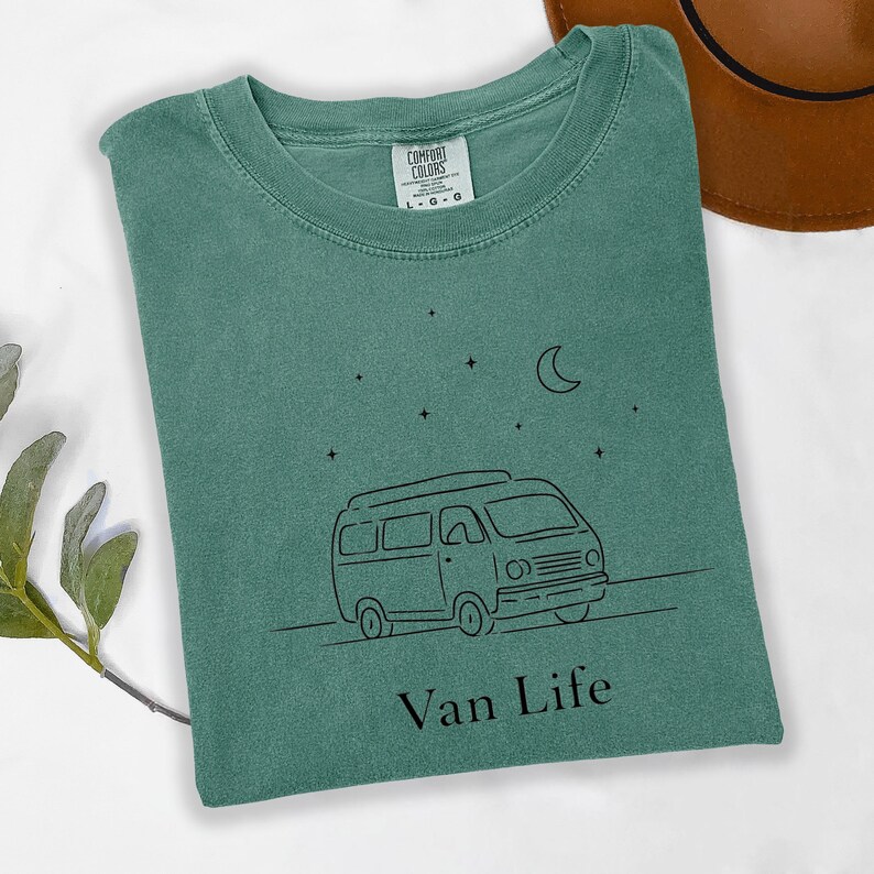 May include: A teal Comfort Colors t-shirt with a black line drawing of a camper van, stars, and a crescent moon. The words "Van Life" are printed below the van. A brown hat is visible in the upper right corner.