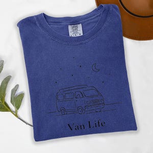 May include: A blue t-shirt with a black line drawing of a van, stars, and a crescent moon. The text "Van Life" is below the van graphic. The shirt has a crew neck and a Comfort Colors tag.