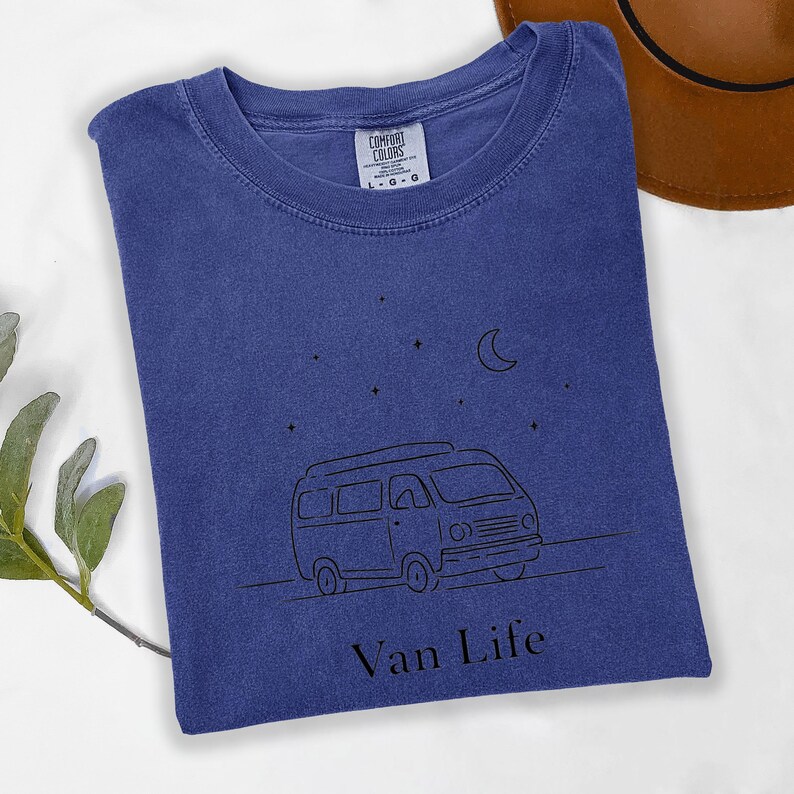 May include: A blue t-shirt with a black line drawing of a van, stars, and a crescent moon. The text "Van Life" is below the van graphic. The shirt has a crew neck and a Comfort Colors tag.