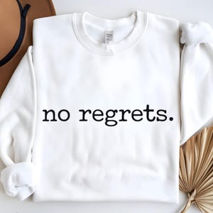 May include: White crewneck sweatshirt with the phrase "no regrets" printed in black. The sweatshirt is made of a soft material and has a classic, casual style. The design is simple and bold, making a statement.