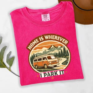 May include: A fuchsia t-shirt with a graphic that reads "HOME IS WHEREVER I PARK IT." The graphic features a vintage orange camper van driving on a road with mountains and trees in the background. The shirt has a crew neck and short sleeves.