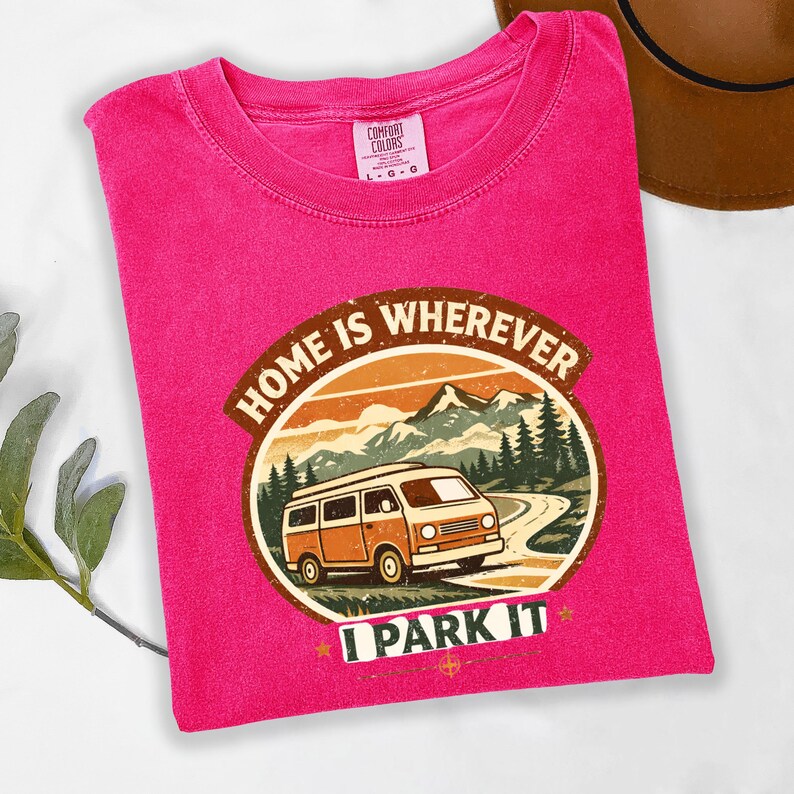 May include: A fuchsia t-shirt with a graphic that reads "HOME IS WHEREVER I PARK IT." The graphic features a vintage orange camper van driving on a road with mountains and trees in the background. The shirt has a crew neck and short sleeves.