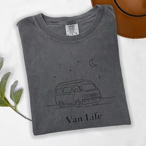 May include: A gray Comfort Colors t-shirt with a black line drawing of a van, stars, and a crescent moon. The text "Van Life" is below the van. A brown hat is partially visible in the upper right corner.