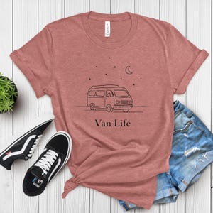 May include: A dusty rose t-shirt with a black line drawing of a van, stars, and a crescent moon. The text "Van Life" is below the graphic. Black and white sneakers and denim shorts are next to the shirt.
