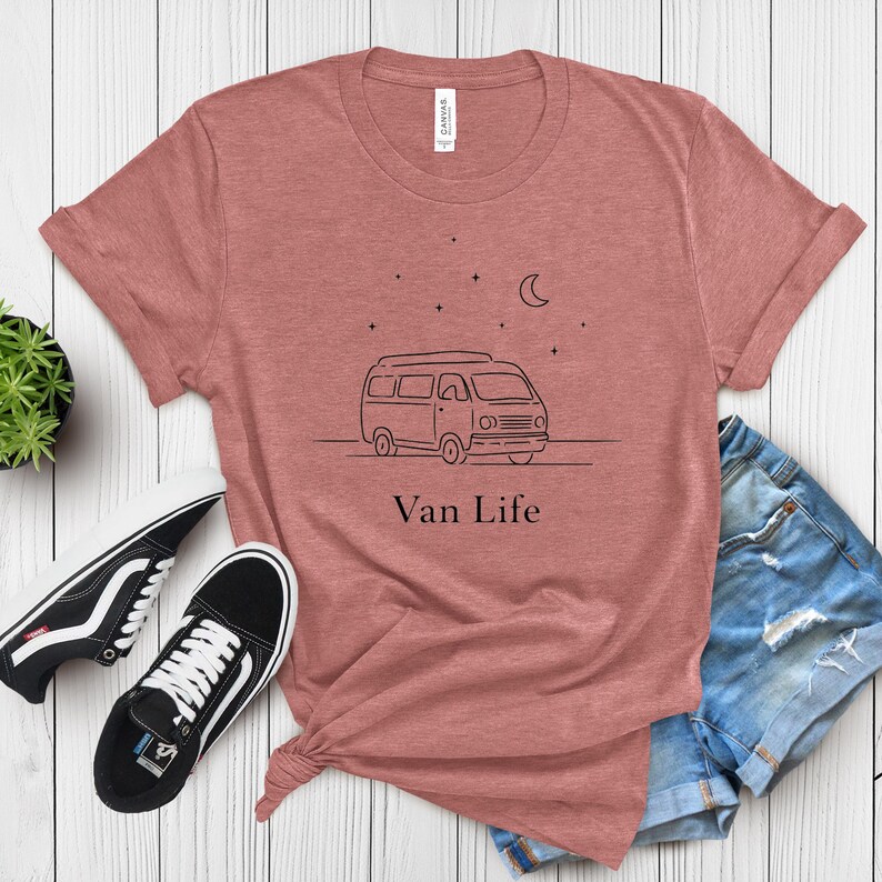 May include: A dusty rose t-shirt with a black line drawing of a van, stars, and a crescent moon. The text "Van Life" is below the graphic. Black and white sneakers and denim shorts are next to the shirt.
