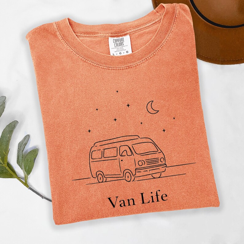 May include: Orange Comfort Colors t-shirt with a black line drawing of a van, stars, and a crescent moon. The text "Van Life" is below the van. The shirt has a crew neck and short sleeves.