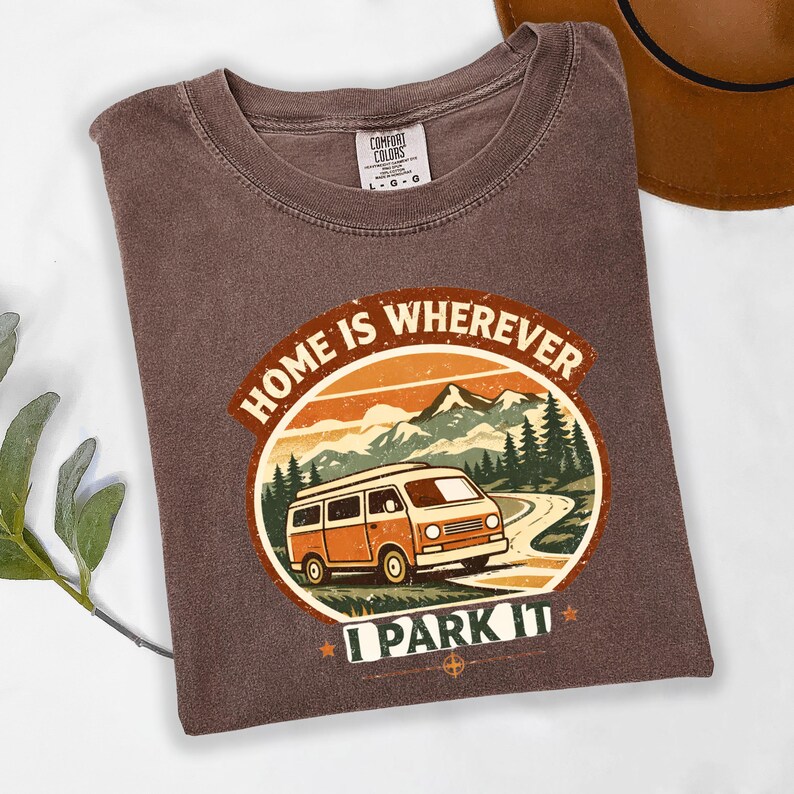 May include: Brown t-shirt with a circular graphic featuring an orange and white camper van driving on a road, with mountains and trees in the background. The text reads "HOME IS WHEREVER I PARK IT."