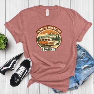 May include: A dusty rose t-shirt with a graphic that reads "HOME IS WHEREVER I PARK IT." The graphic features an orange camper van driving on a road with mountains and trees in the background. Black and white sneakers and denim shorts are next to the shirt.