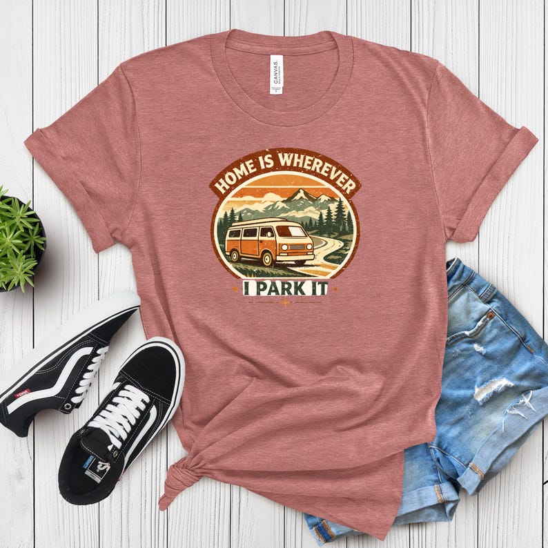 May include: A dusty rose t-shirt with a graphic that reads "HOME IS WHEREVER I PARK IT." The graphic features an orange camper van driving on a road with mountains and trees in the background. Black and white sneakers and denim shorts are next to the shirt.