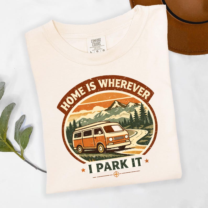 May include: Cream-colored t-shirt with a circular graphic featuring an orange and white van driving on a winding road through a mountain landscape. The text "HOME IS WHEREVER I PARK IT" is displayed in a brown and orange design.