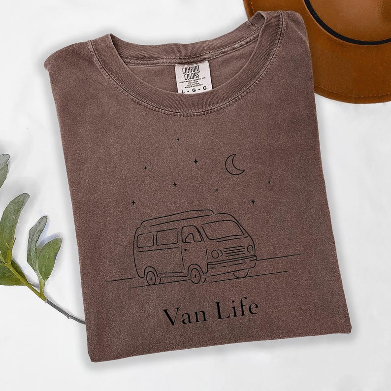 May include: A brown t-shirt with a black line drawing of a camper van, stars, and a crescent moon. The text "Van Life" is below the van. The shirt is a Comfort Colors brand, size L-G.