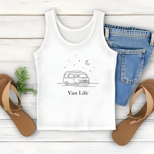 May include: White tank top with a black line drawing of a van, stars, and a crescent moon. The text "Van Life" is below the drawing. The tank top is paired with blue jeans and brown sandals.