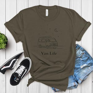 May include: Olive green t-shirt with a black line drawing of a van, stars, and a crescent moon. The text "Van Life" is below the drawing. Black and white sneakers and denim shorts are also in the image.
