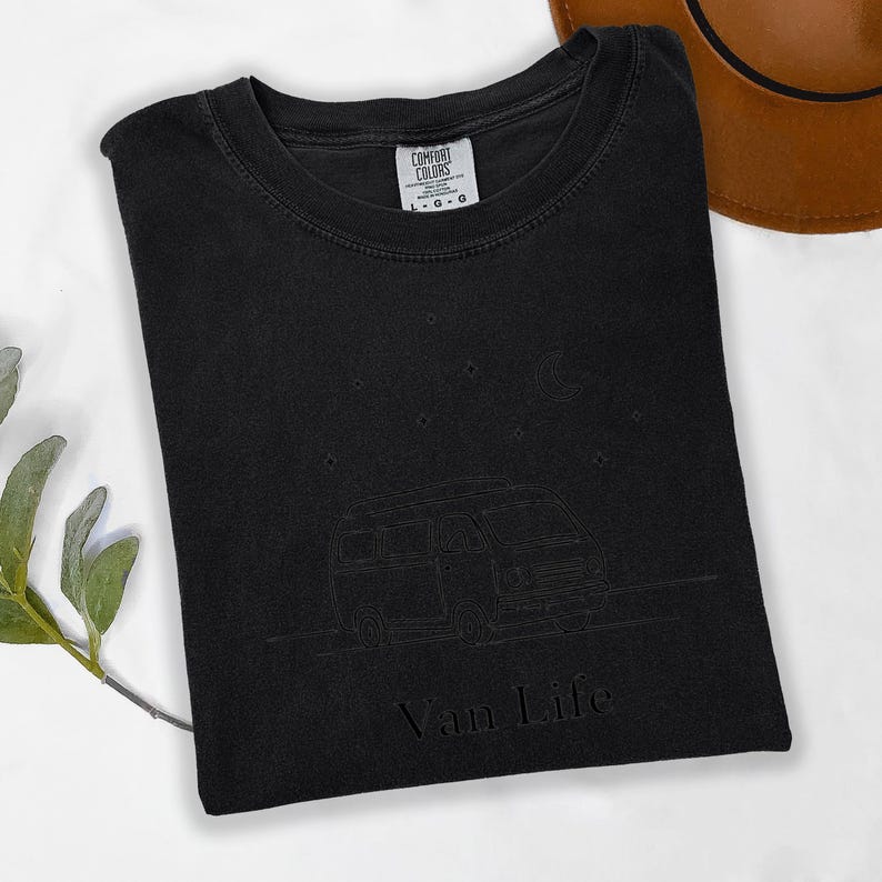 May include: Black t-shirt with a line drawing of a camper van under a night sky with stars and a crescent moon. The words "Van Life" are below the van. The shirt is folded on a white surface.