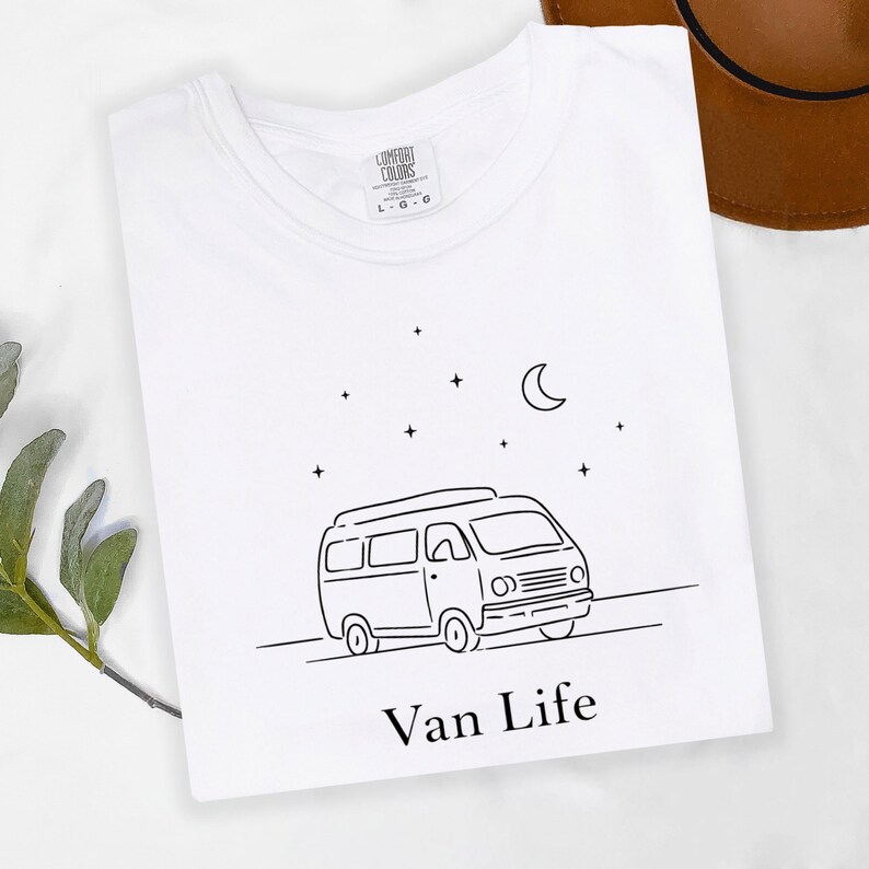 May include: White t-shirt with a black line drawing of a camper van, stars, and a crescent moon. The text "Van Life" is below the van. The shirt has a crew neck and a tag that says "Comfort Colors".