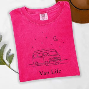 May include: A hot pink t-shirt with a crew neck. The shirt features a black line drawing of a van, stars, and a crescent moon. The words "Van Life" are printed below the van.
