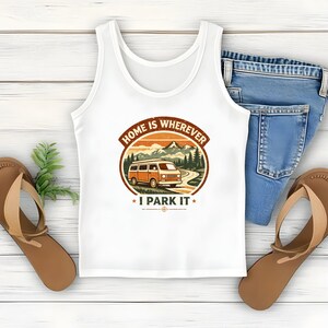 May include: White tank top with a graphic that reads "HOME IS WHEREVER I PARK IT" featuring an orange camper van, mountains, and trees. The tank top is paired with blue jeans and brown sandals.