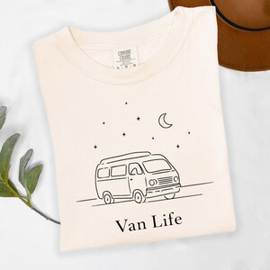 May include: Cream-colored t-shirt with a black line drawing of a camper van, a crescent moon, and stars. The words "Van Life" are printed below the van. A brown hat is visible in the background.