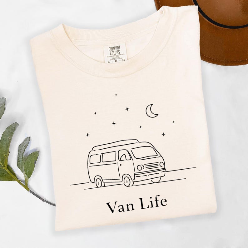 May include: Cream-colored t-shirt with a black line drawing of a camper van, a crescent moon, and stars. The words "Van Life" are printed below the van. A brown hat is visible in the background.