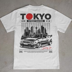 May include: White t-shirt with a graphic of a black and silver sports car set against a grey cityscape. The text "TOKYO STREET RACING" is above the car, with Japanese characters. The words "Shooting Star" are also present.