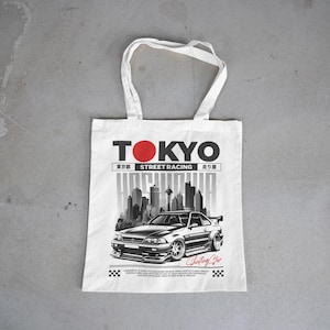 May include: White tote bag with a graphic design featuring the word "TOKYO" in black with a red circle, a cityscape, and a black and silver car. The words "Street Racing" are also included.