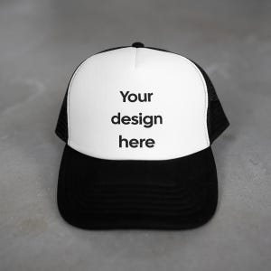 May include: A black and white trucker cap with a white front panel. The front panel displays the text "Your design here" in black. The cap has a black peak and mesh back.