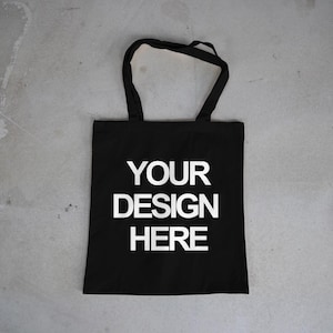 May include: Black tote bag with the text "YOUR DESIGN HERE" in white. The bag has two black handles and is made of a solid black material. The bag is designed to be customized.