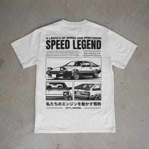 May include: White t-shirt featuring a black car graphic and text. The text reads "A LEGACY OF SPEED AND PRECISION SPEED LEGEND". Below the text are three illustrations of the car. The bottom of the shirt has Japanese text and the brand name.