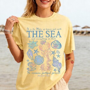 May include: A pale yellow short-sleeve t-shirt featuring a blue graphic print of seashells, a lobster, and a crab. The text reads: He is the Maker of heaven and earth THE SEA and everything in them He remains faithful forever. PSALM 146:6.