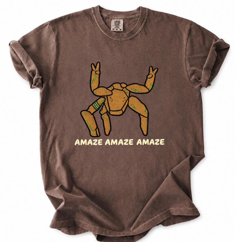 May include: A brown short-sleeve t-shirt featuring a graphic of a tan, multi-legged creature with green accents making peace signs with its top limbs. The text "AMAZE AMAZE AMAZE" is printed in light yellow capital letters below the creature graphic.