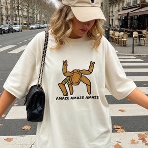 May include: An off-white short-sleeve t-shirt featuring a graphic of a tan, multi-legged creature with green accents making peace signs with two upper limbs. The text "AMAZE AMAZE AMAZE" is printed in black capital letters below the graphic.