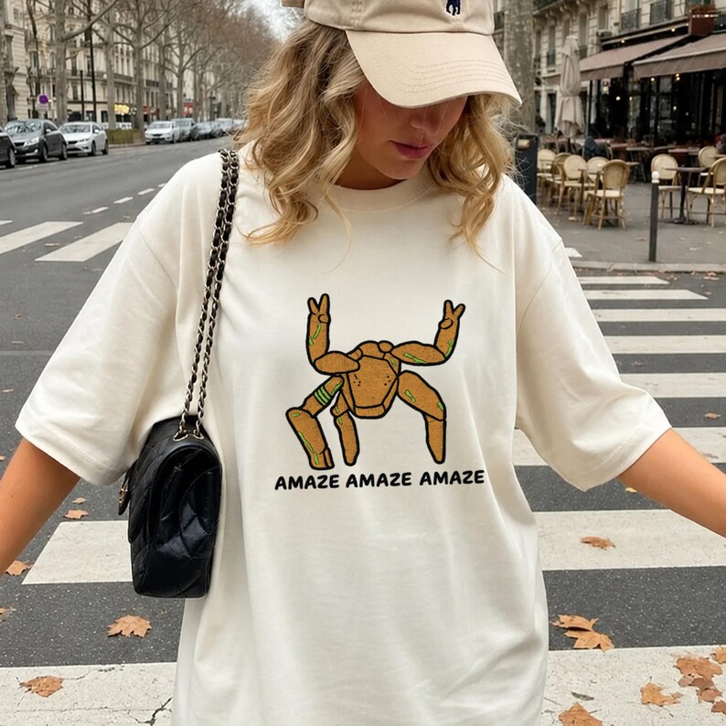 May include: An off-white short-sleeve t-shirt featuring a graphic of a tan, multi-legged creature with green accents making peace signs with two upper limbs. The text "AMAZE AMAZE AMAZE" is printed in black capital letters below the graphic.