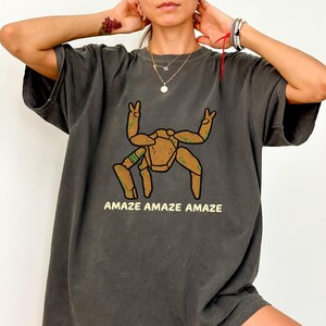 May include: A charcoal gray short-sleeve t-shirt featuring a graphic of a brown, multi-legged creature with green accents making peace signs with its top limbs. The text "AMAZE AMAZE AMAZE" is printed in a light yellow font below the graphic.
