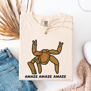 May include: A cream-colored short-sleeve t-shirt featuring a brown, wood-textured creature graphic with green accents. The creature has four legs and two arms raised in peace signs. The text "AMAZE AMAZE AMAZE" is printed in black below the graphic.