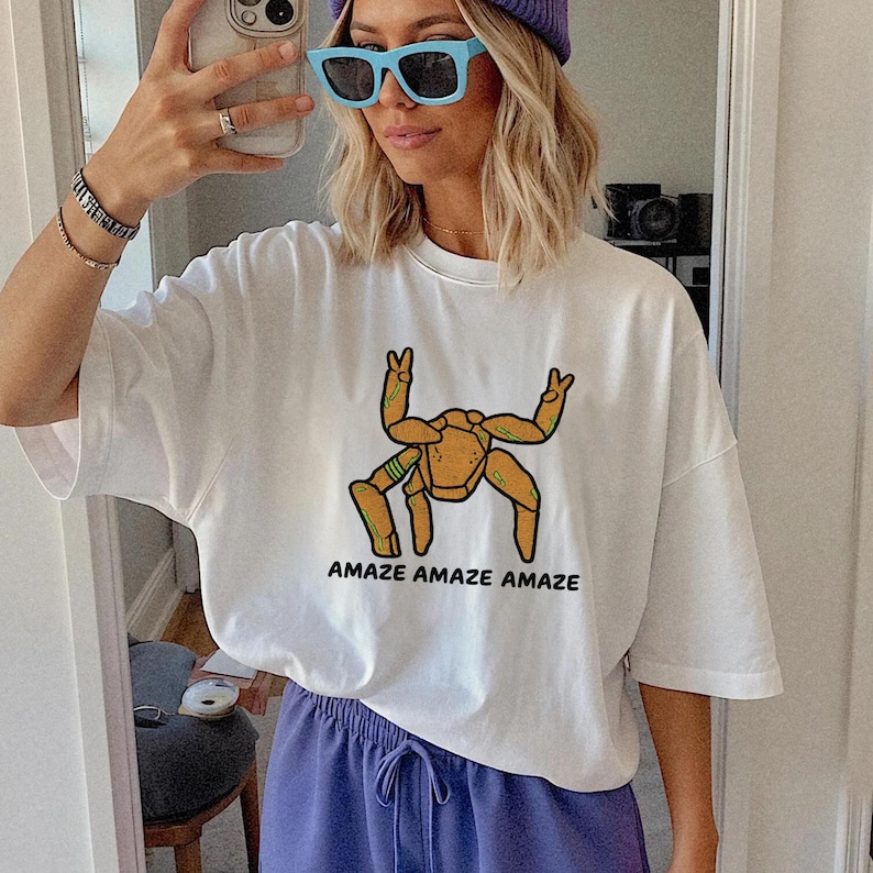 May include: A white short-sleeved t-shirt featuring a graphic of a tan, multi-legged creature with green accents making peace signs with its upper limbs. Below the graphic, the text reads AMAZE AMAZE AMAZE in bold, black, sans-serif capital letters.