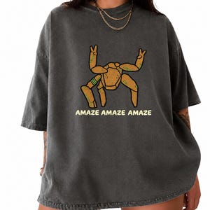 May include: A charcoal gray short-sleeve t-shirt featuring a graphic of a tan, multi-legged creature with green accents making peace signs with its top limbs. The text "AMAZE AMAZE AMAZE" is printed in a light yellow font below the creature illustration.