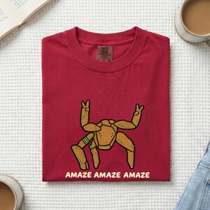 May include: A dark red t-shirt featuring a graphic of a tan, multi-legged creature with green accents making peace signs with two of its limbs. The text "AMAZE AMAZE AMAZE" is printed in a light yellow font below the graphic on the front of the shirt.